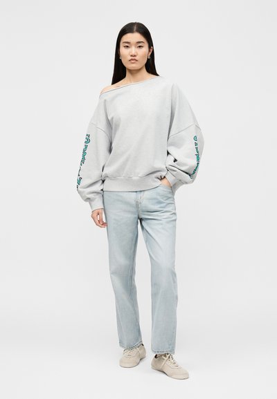 Νέα γυναίκα με μακριά μαύρα μαλλιά που φοράει φαρδιά ανοιχτογκρι φούτερ χωρίς ώμους και ανοιχτό μπλε τζιν, στέκεται με το ένα χέρι στην τσέπη.