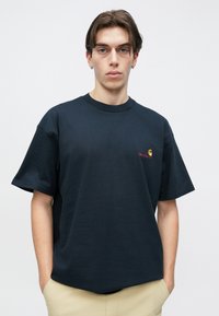 Σκούρο μπλε βαμβακερό T-shirt, κοντά μανίκια, στρογγυλή λαιμόκοψη, με μικρό γραφικό λογότυπο σε κίτρινο και κόκκινο στην αριστερή πλευρά του στήθους.