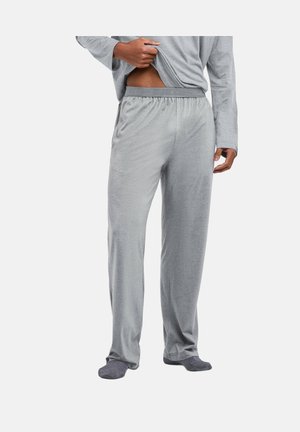 Man met losse grijze pyjamabroek en bijpassende top met lange mouwen, hij tilt zijn overhemd op om de tailleband te laten zien, staat blootsvoets op een witte achtergrond.