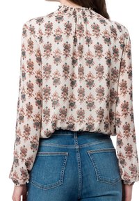 Femme portant une blouse crème à manches longues avec un motif floral marron et turquoise, rentrée dans un jean en denim bleu, vue de dos.