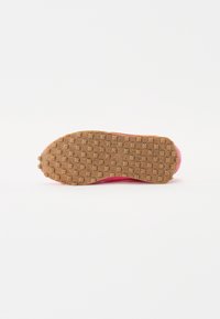 Semelle d'une chaussure de sport rose avec une semelle en caoutchouc marron texturée comportant de petites gommes carrées sur fond blanc.