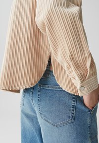 Maglietta beige a maniche lunghe con orlo arrotondato, con polsini a bottoni e fessure laterali. Indossata sopra jeans di denim blu chiaro.