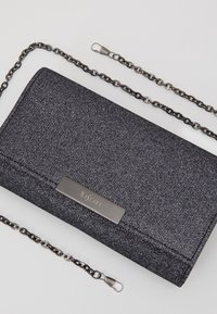 Portefeuille texturé noir avec une plaque logo argentée, équipé d'une bandoulière chaîne pour un port en bandoulière. Forme rectangulaire au design élégant.