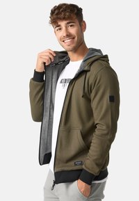 Olivgrüner Reißverschluss-Hoodie mit grauem Futter, Kordelzugkapuze, schwarzen Akzenten und zwei vorderen Taschen. Weicher, strukturiertes Material.