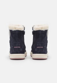 Bottines en suède marine avec doublure en fourrure crème, présentant un logo rose pâle et une semelle en caoutchouc contrastante. Conçues avec un bout arrondi et une fermeture latérale.