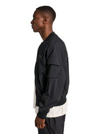 Junger Mann im Profil, der eine schwarze Bomberjacke über einem weißen Hemd trägt, mit schwarzen Hosen und einem Creolen-Ohrring vor weißem Hintergrund.