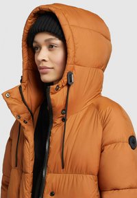 Polstret orange jakke med en stor hætte, featuring sorte accenter, justerbare snore og en frontlynlås. Båret med en sort beanie.