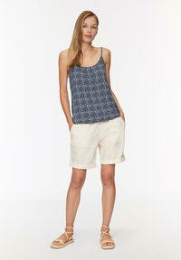 Mouwloos navy tanktop met een geometrisch wit patroon, gecombineerd met crèmekleurige opgerolde shorts. Model staat met de handen in de zakken en draagt beige sandalen.