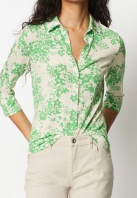 Grön och vit blommönstrad blus med knappstängning fram och trekvartslånga ärmar, tillverkad av lätt tyg. Matchad med ljusbeiga byxor.