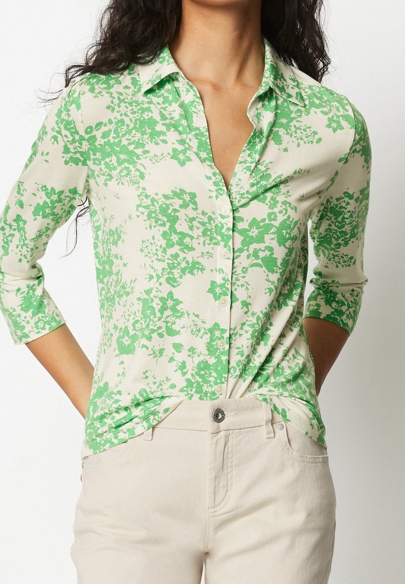 Grön och vit blommönstrad blus med knappstängning fram och trekvartslånga ärmar, tillverkad av lätt tyg. Matchad med ljusbeiga byxor.