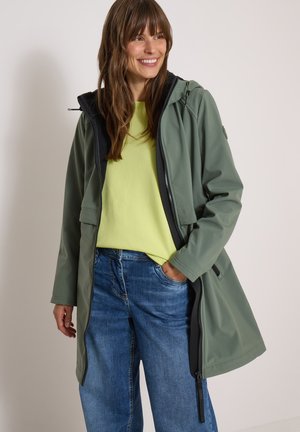 Smilende kvinde iført grøn jakke over gul skjorte og blå jeans, stående med den ene hånd i lommen mod ensfarvet baggrund.