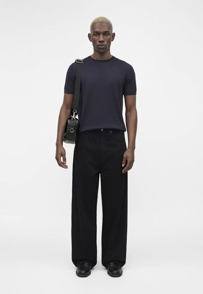 Νέος άνδρας με κοντά ξανθά μαλλιά φορώντας μαύρο παντελόνι με φαρδιά μπατζάκια, σκούρο μπλε μπλουζάκι, μαύρα παπούτσια και μαύρη τσάντα ώμου.