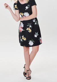 Robe fleurie noire ornée de fleurs blanches, jaunes et roses, manches courtes et taille cintrée. Associée à des talons noirs à brides.