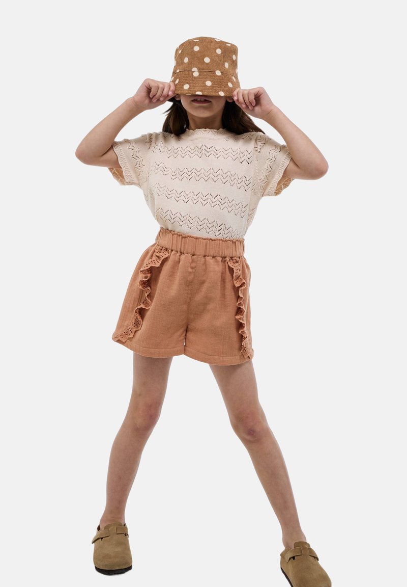 Chapeau seau tan à pois blancs, haut en tricot crème avec un motif zigzag, et shorts rouille avec des côtés volantés. Chaussures brunes à enfiler.
