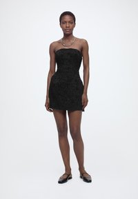 Γυναίκα όρθια, φοράει ένα μαύρο μίνι φόρεμα χωρίς τιράντες με υφή, μαύρα επίπεδα παπούτσια και χοντρό ασημένιο κολιέ σε λευκό φόντο.