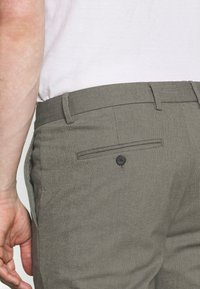 Pantalones grises texturizados con un bolsillo trasero abotonado. Presentan una cinturilla con passadores para cinturón y un pliegue cosido en la parte delantera.