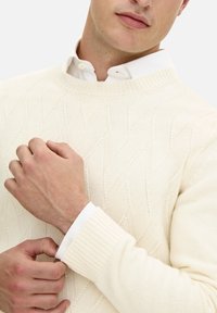 Maglione lavorato a maglia color crema con motivo a trecce, caratterizzato da un collo a girocollo a coste e maniche lunghe sovrapposte a una camicia bianca con colletto.