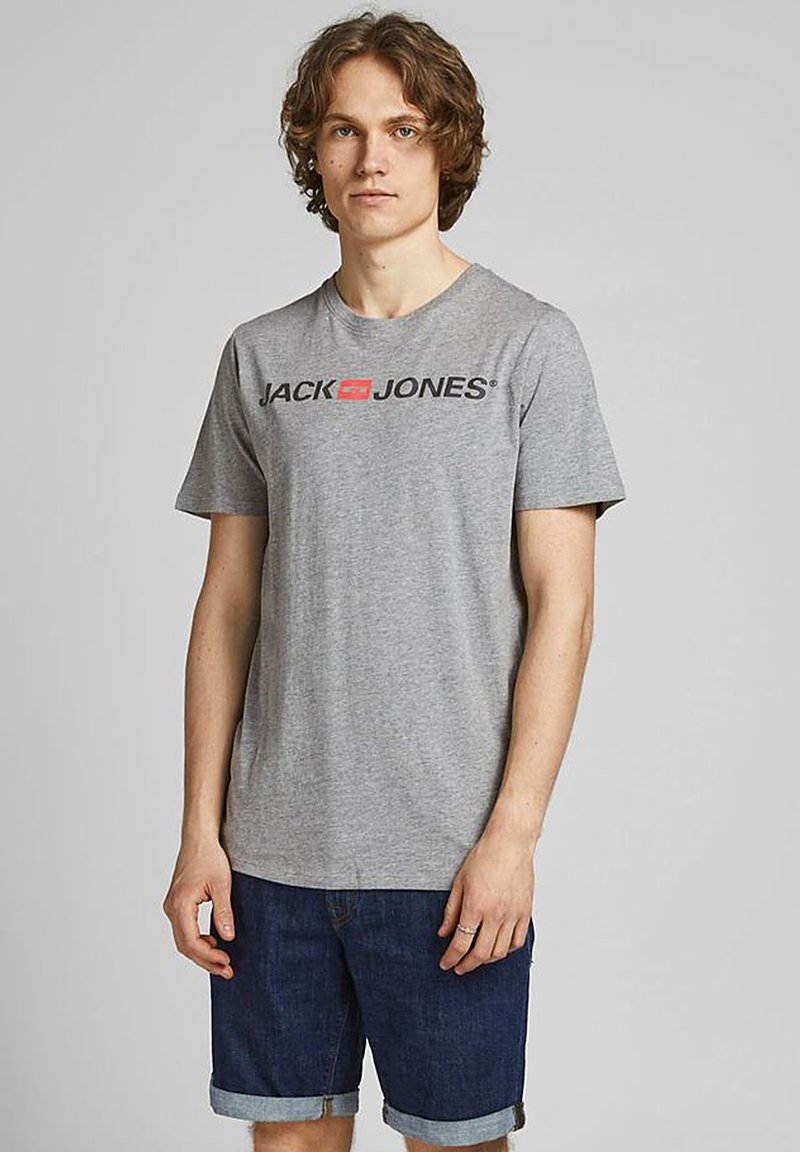 Graues Baumwoll-T-Shirt mit kurzen Ärmeln, versehen mit einem schwarzen und roten "JACK & JONES"-Logo auf der Brust. Trägt dazu eine Jeansshorts.