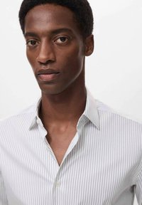 Camisa blanca de botones a rayas con un cuello clásico, textura suave y líneas verticales sutiles, llevada por una persona con pelo corto.