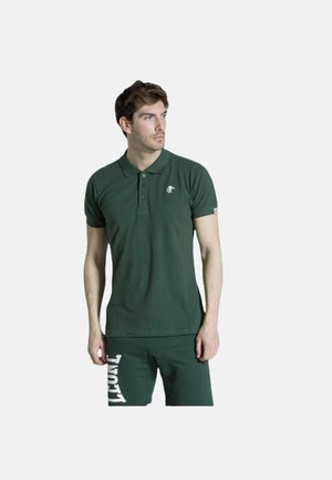 Giovane uomo che indossa una polo verde scuro e pantaloncini abbinati con testo bianco, in piedi davanti a uno sfondo chiaro uniforme, guardando a destra.