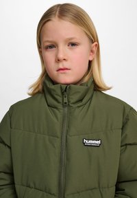 Grön puffjacka med hög krage, framdragkedja och "hummel"-logotyp. Har ett vadderat mönster och ett slitstarkt tyg.