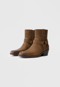 Botas de cuero marrón hasta el tobillo con una textura suave, cremallera lateral y una correa decorativa con acentos dorados; tacón bajo y ancho.