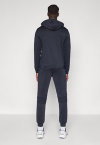 Tumši zils kapučjakas un bikses komplekts ar fakturētu dizainu un ribveida aprocēm, uz kapučjaka aizmugures izšūts logotips.