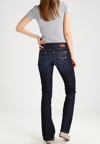 Vrouw in donkerblauwe bootcut jeans en een grijs shirt met korte mouwen, staand met één hand op haar heup, met de rug naar de camera op een effen achtergrond.