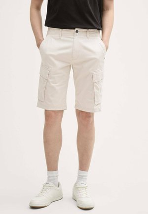 Beige Cargo-Shorts mit Seitentaschen, Knopfverschluss und Gürtelschlaufen. Kombiniert mit einem schwarzen T-Shirt und weißen Sneakers. Aus Baumwollmaterial.