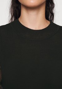 Top en maille côtelée noire avec un col rond et un tissu texturé. La coupe est ajustée, avec des détails de surpiqûres visibles autour du col.