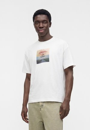 Jeune homme portant un t-shirt blanc avec un motif de lèvres, d'étoiles et le texte "No Signal", associé à un pantalon beige clair sur un fond uni.
