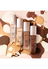 Quatre bouteilles de fond de teint soin Flawless Finish d'Elizabeth Arden dans des teintes variées, posées sur des tourbillons de fond de teint crème et marron.