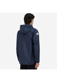 Marinblå vattentät jacka med huva, har elastiska ärmslut och en logotyp på höger axel, slät textur och avslappnad passform.