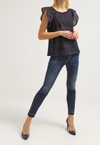 Haut bleu marine avec détails en dentelle et manches volantées, associé à un jean foncé ajusté et des talons pointus. Tissu texturé sur toute la surface.