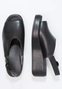 Černé kožené skluzné boty s otevřenou špičkou, silnou platformou a nastavitelným páskem kolem paty. Obsahují kulatý kovový detail.