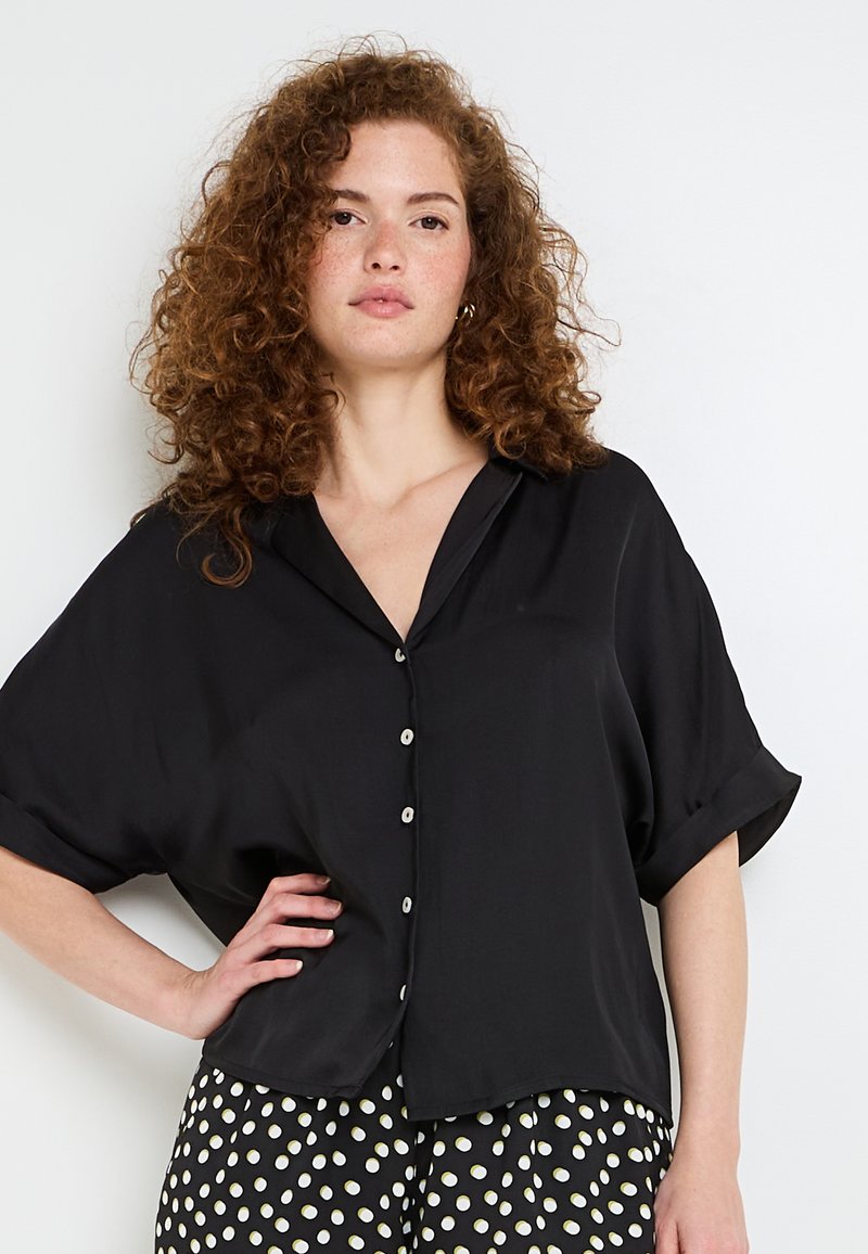 Chemise noire à manches courtes avec un fit décontracté. Texture lisse, ourlet légèrement incurvé et col cranté. Les boutons sont de couleur claire.
