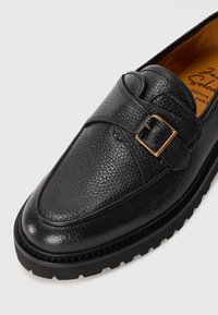 Black textured leather shoe with a rounded toe, featuring a side buckle accent and a durable rubber sole. Interior shows brand text.
