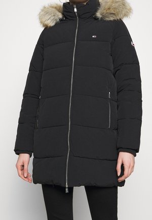 Abrigo negro acolchado de invierno con capucha forrada de piel, cremallera frontal y bolsillos laterales con cremallera, llevado por una persona de pie.