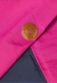 Růžová tkanina s hladkou strukturou, na které je kulatá hnědá kožená nášivka s vyraženým logem a geometrickým designem, umístěná na tmavě šedém pozadí.