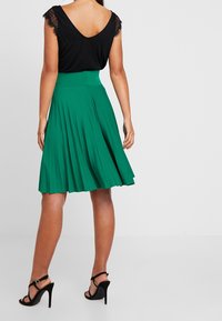 Top nero con maniche in pizzo, abbinato a una gonna plissettata verde fino al ginocchio. Sandali con tacco alto neri completano il look.