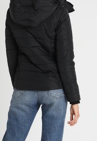 Svart pufferjacka med huva, med en quiltad textur och en slim fit. Matchas med högmidjade blå jeans, vilket visar en avslappnad stil.