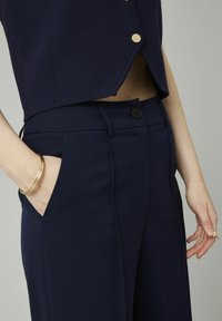Pantalons ajustés bleu marine avec une texture lisse, dotés d'une fermeture par bouton, de poches inclinées et d'une coupe structurée. Bracelet doré visible.