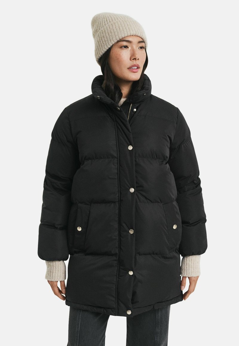 Schwarze Steppjacke mit großen, gesteppten Abschnitten, silbernen Druckknöpfen und seitlichen Taschen. Sie hat einen hohen Kragen und einen dicken Stoff für Wärme.