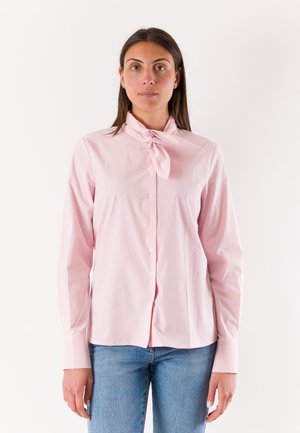 Donna con lunghi capelli scuri indossa una camicetta rosa chiaro con bottoni e fiocco al collo e jeans blu, in piedi su sfondo bianco semplice.