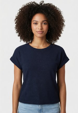 Giovane donna con capelli ricci che indossa una maglietta blu navy a maniche corte e jeans azzurri, rivolta verso l'obiettivo su uno sfondo chiaro e uniforme.