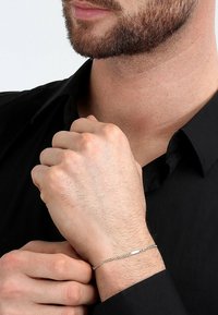 Silberne Kettenarmband an einem Handgelenk, mit ineinandergreifenden Gliedern und einem flachen Stabdesign. Der Träger hat einen hellen Hautton und einen kurzen Bart.