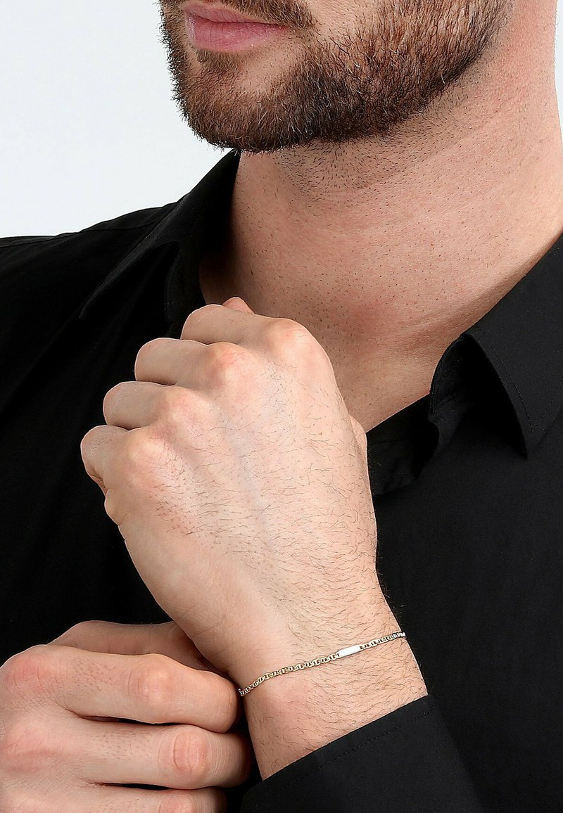 Silberne Kettenarmband an einem Handgelenk, mit ineinandergreifenden Gliedern und einem flachen Stabdesign. Der Träger hat einen hellen Hautton und einen kurzen Bart.