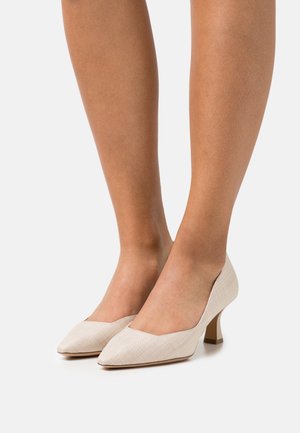 Jambes portant des escarpins à talons bas pointus beige texturé sur un fond blanc.