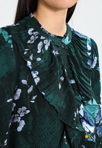 Blusa verde oscura con patrón floral, volantes plisados en el cuello y botones negros. Tela ligera con textura suave.