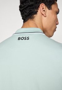 Svetlomodré polo tričko s čiernym vyšívaným logom "BOSS" na chrbte, so kontrastným lemovaným golierom a textúrovanou látkou.